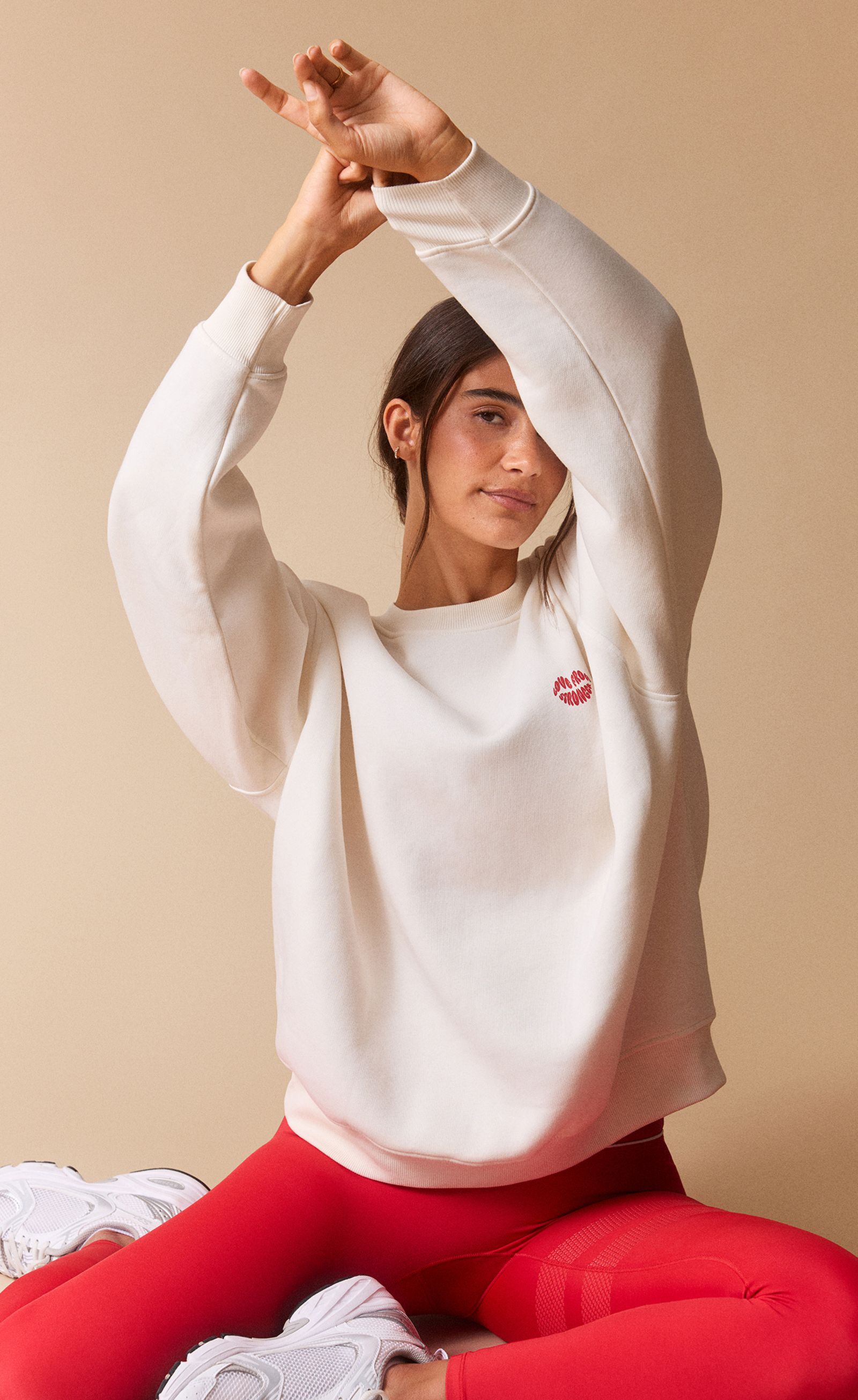 Person sitting cross-legged, wearing a cream sweatshirt with red heart detail and bright red pants, against a beige background.