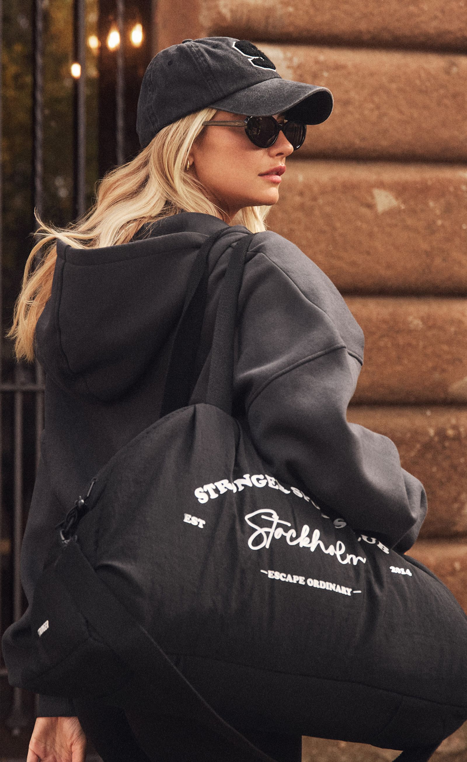 Blonde woman in black cap, sunglasses and hoodie, carrying a black duffel bag reading "Stockholm" in front of a stone wall.