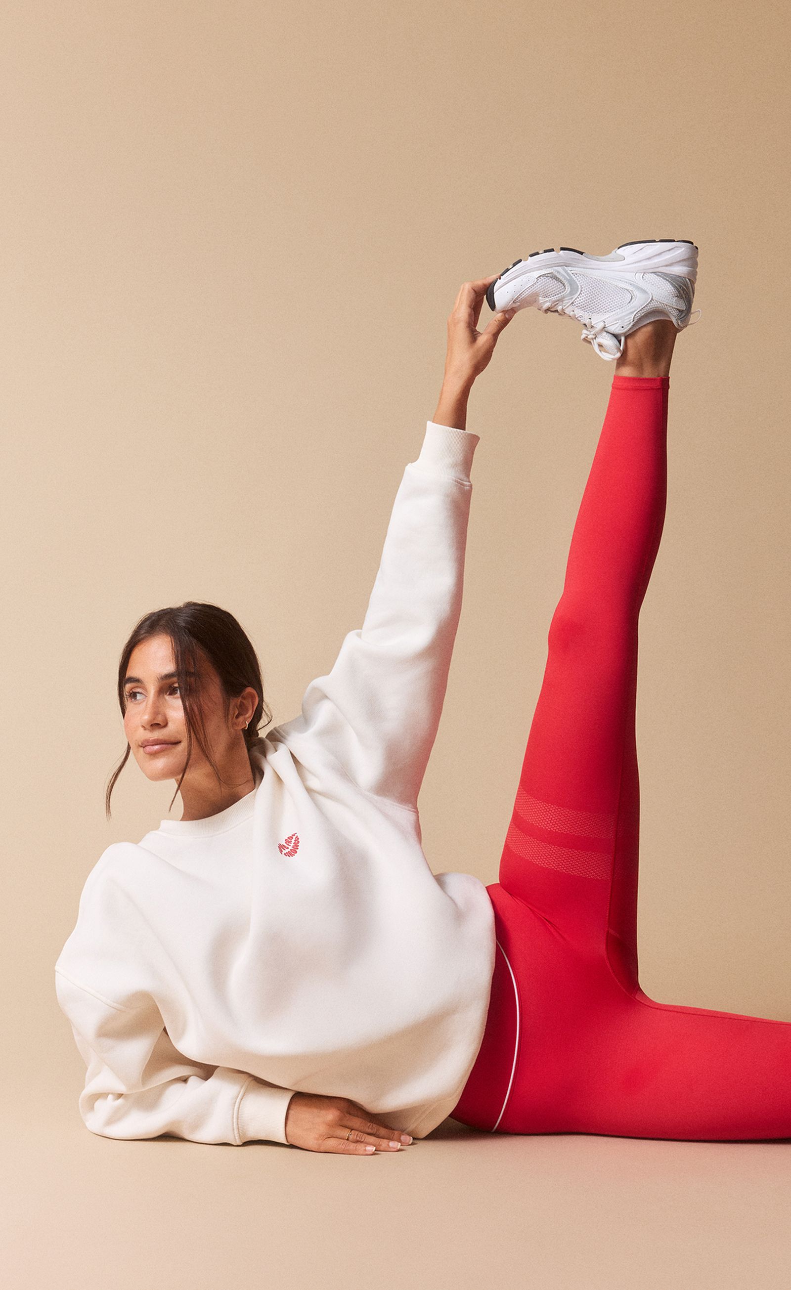 A woman in a white sweatshirt and red leggings stretches on the floor, lifting one leg high while smiling and looking upward.
