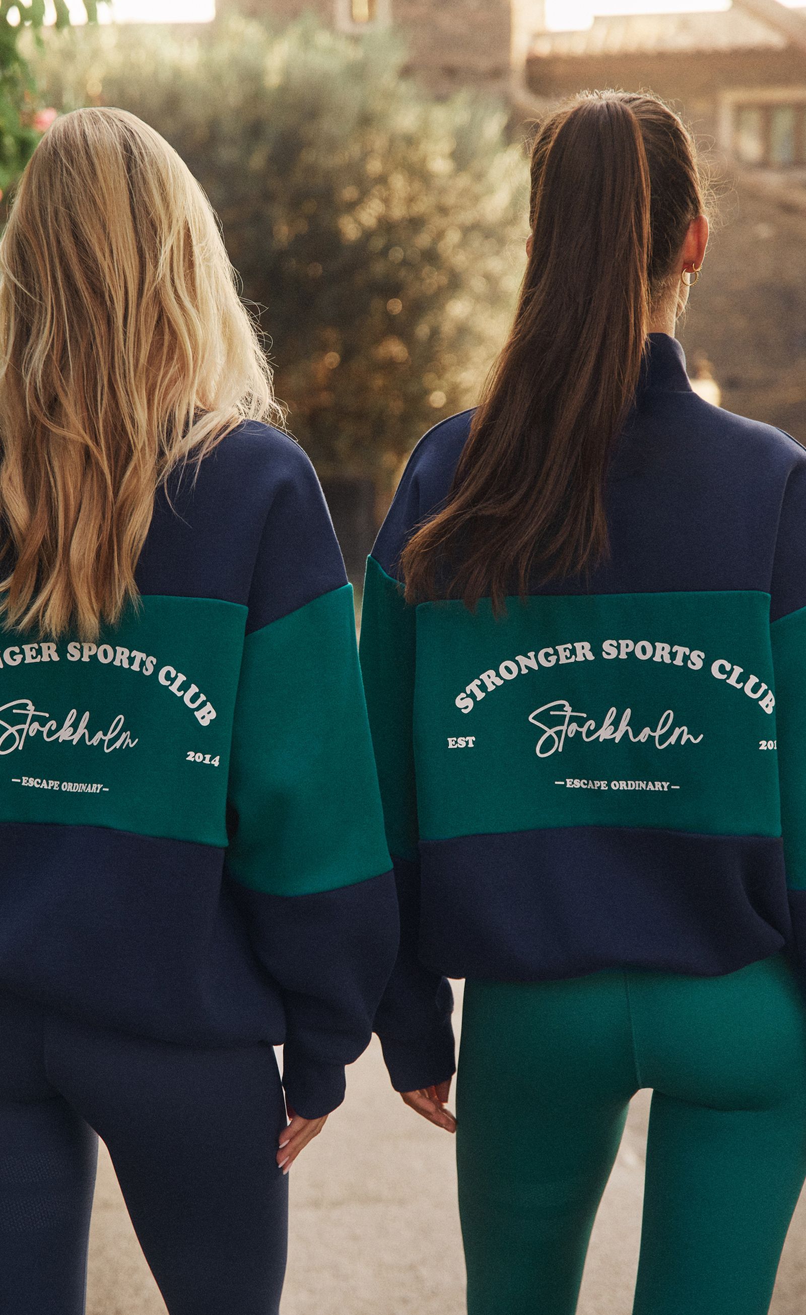 Two women wearing "Stronger Sports Club Stockholm" sweatshirts and leggings walk outdoors, one with blonde hair and the other with a ponytail.