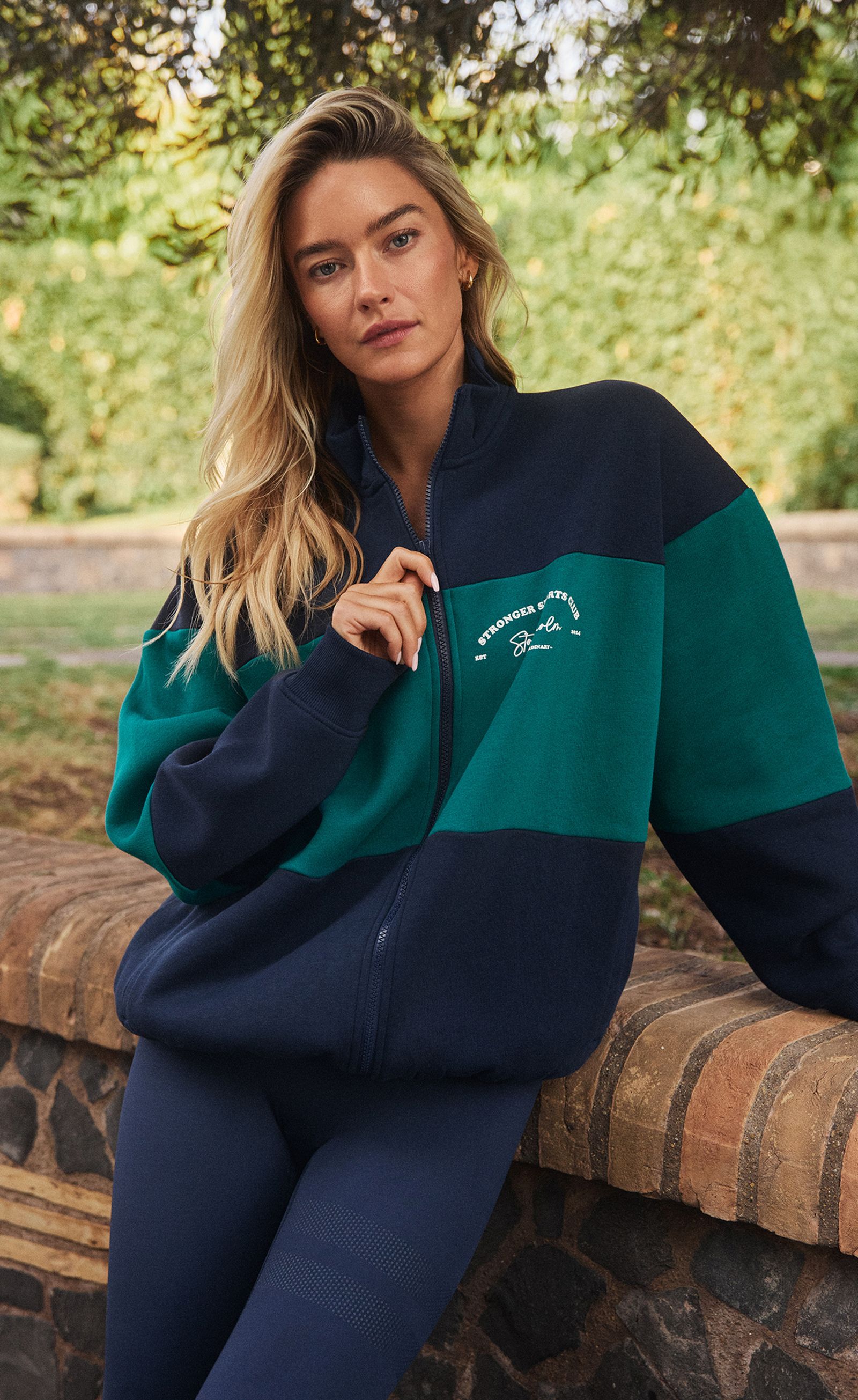 Blonde woman with loose hair leans on a brick wall outdoors, wearing a navy-and-teal zip-up jacket and navy leggings, neutral expression.