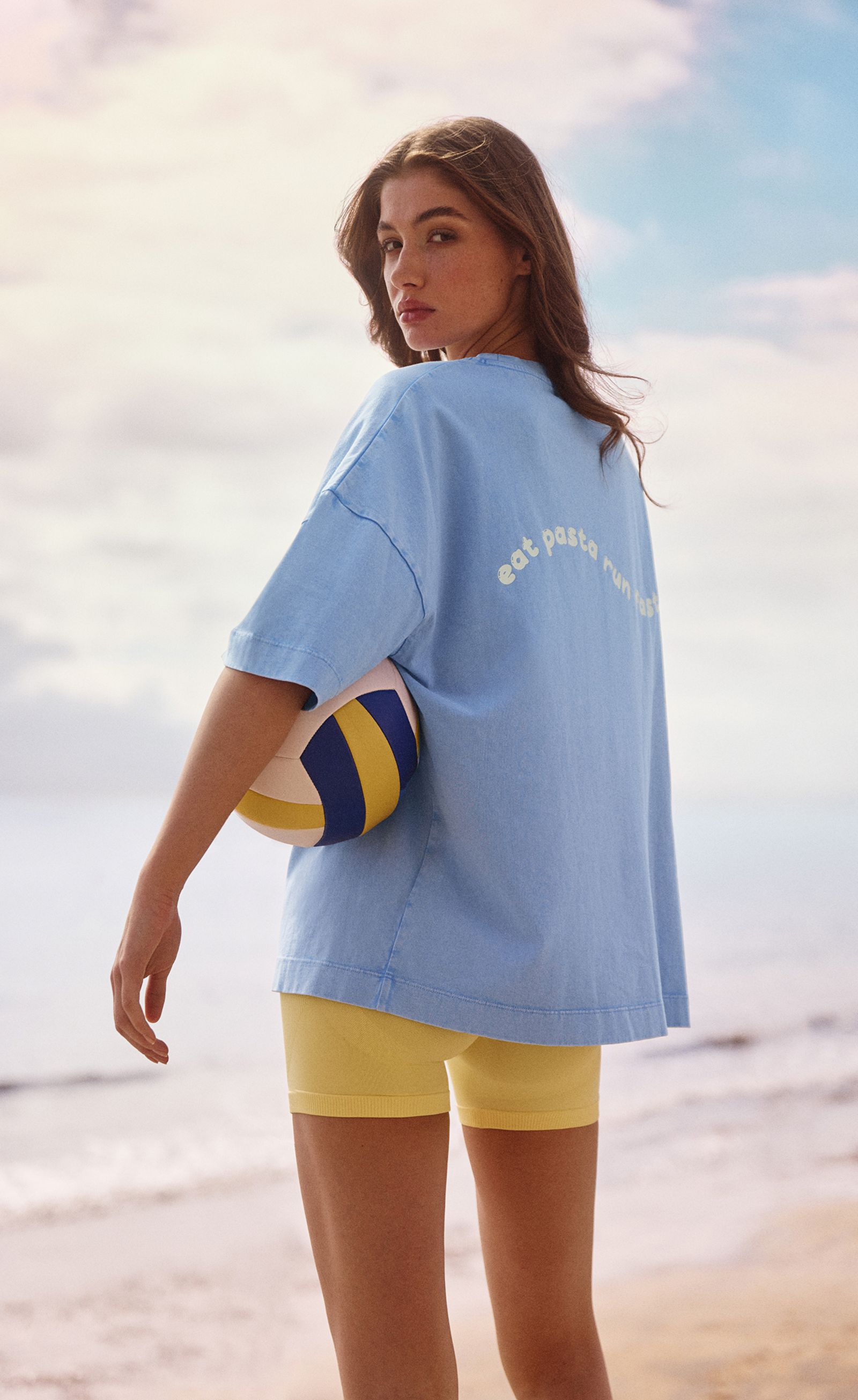 A woman on the beach wearing a blue shirt and yellow shorts, holding a volleyball, with the ocean and sky in the background.