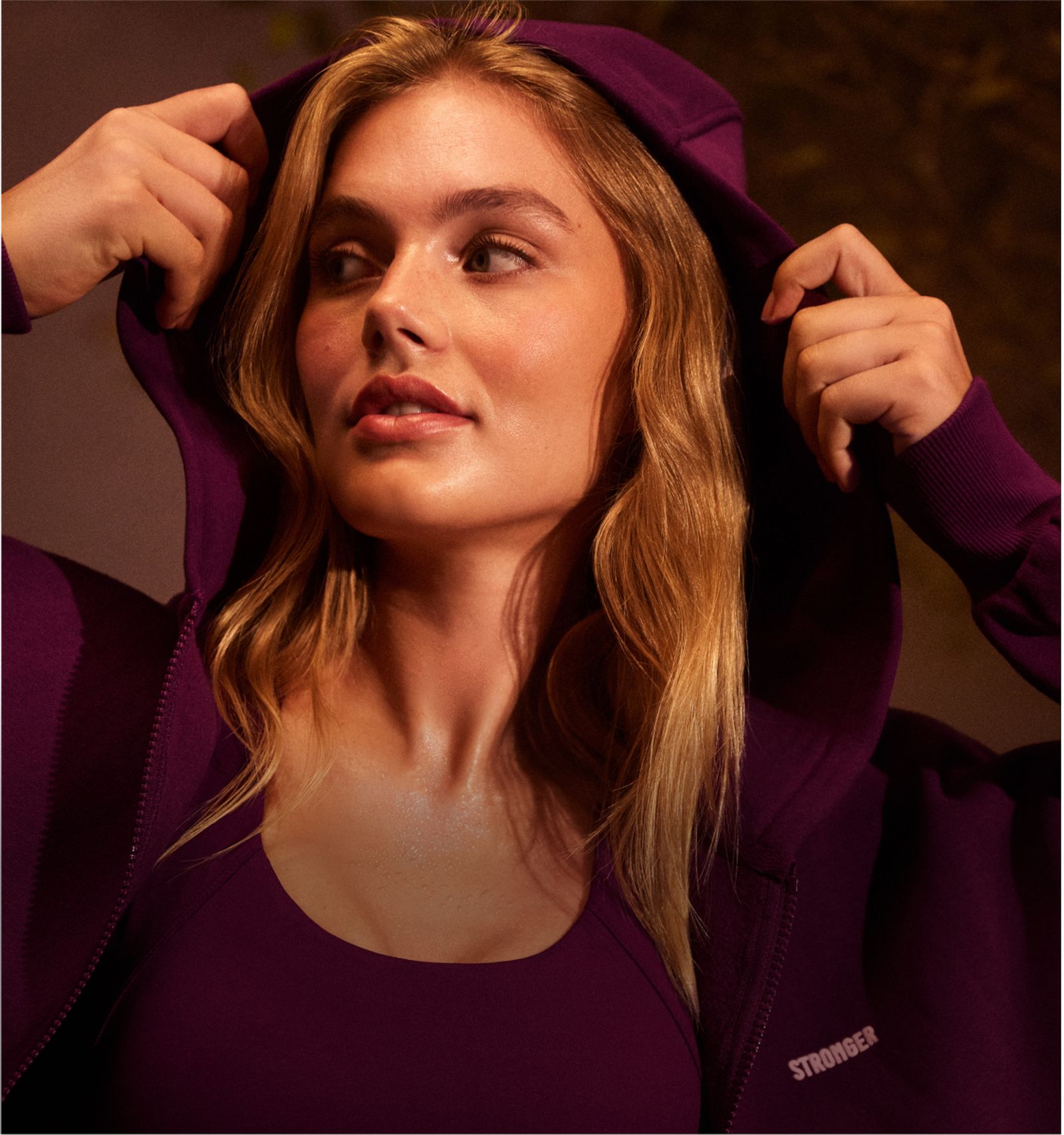 Person with long hair wearing a purple hoodie and tank top, looking to the side, adjusting the hood with both hands.