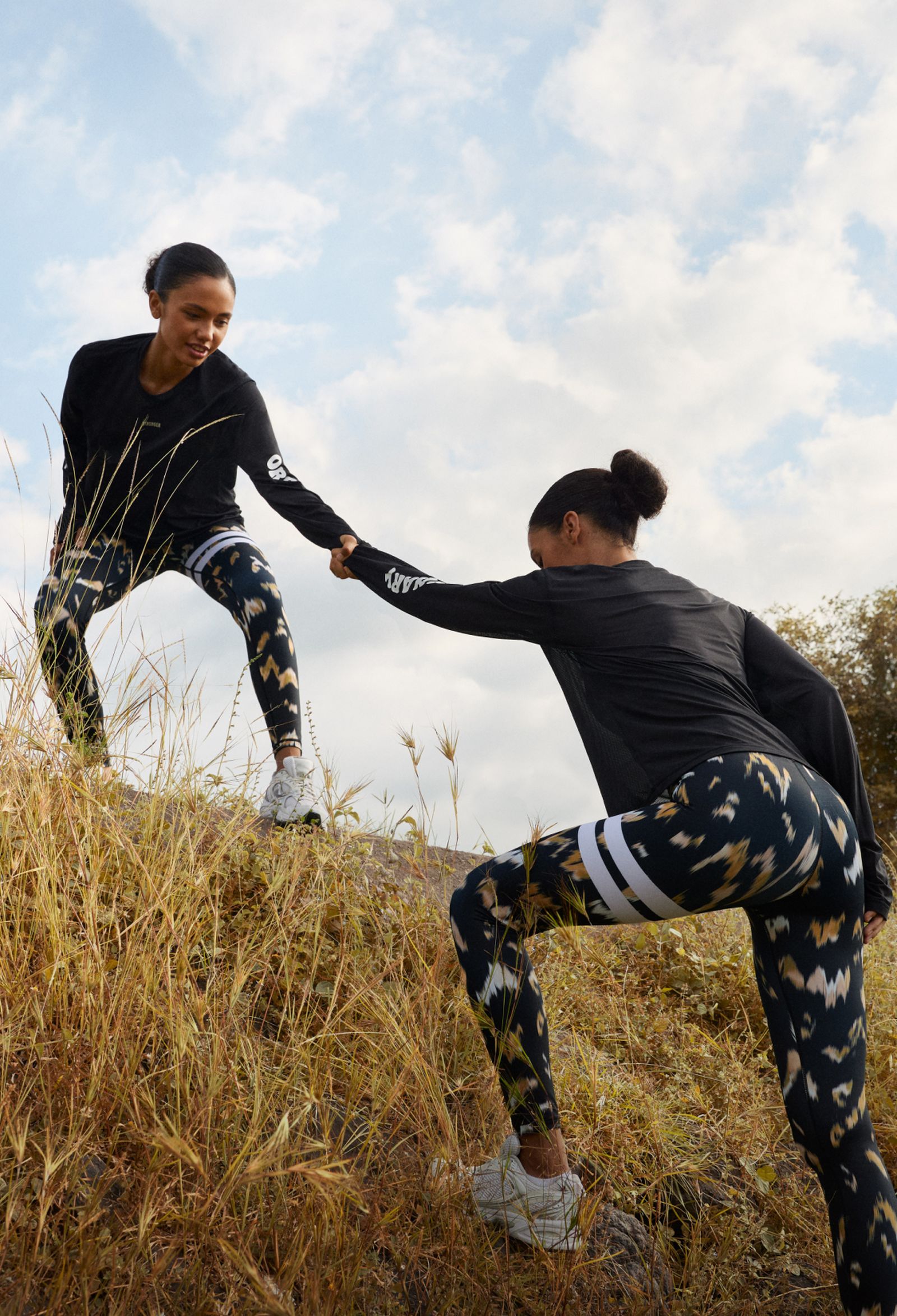 Two women in athletic wear on a grassy slope, one helping the other up, with a cloudy sky in the background.