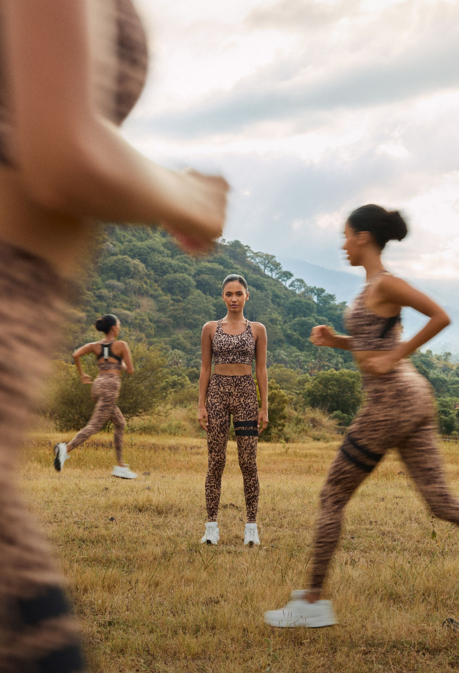 Woman in leopard print workout attire stands in a field as blurred figures run past, with green hills in the background.
