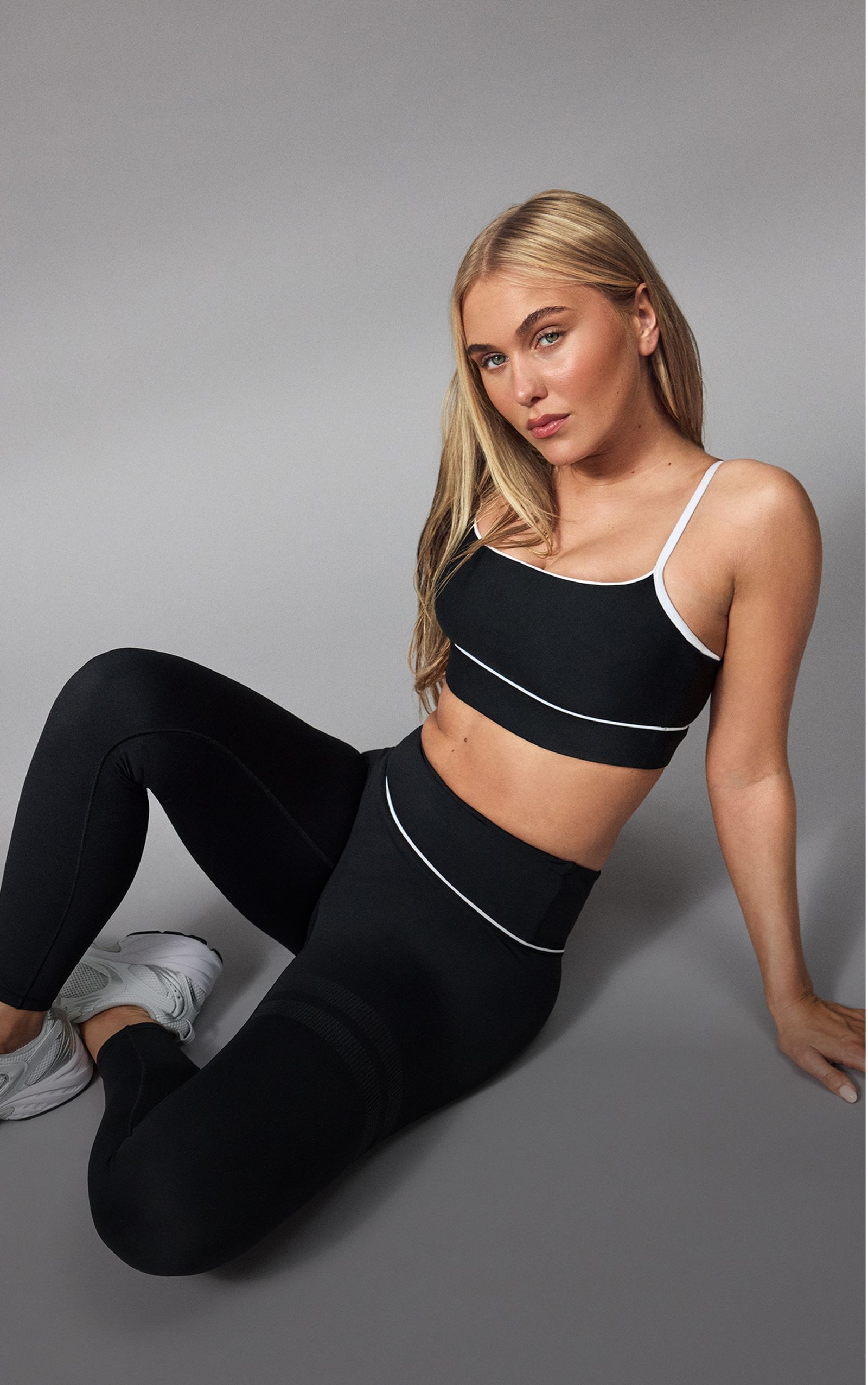 Woman in black workout attire with white trim sitting on the floor, posing against a neutral gray background.