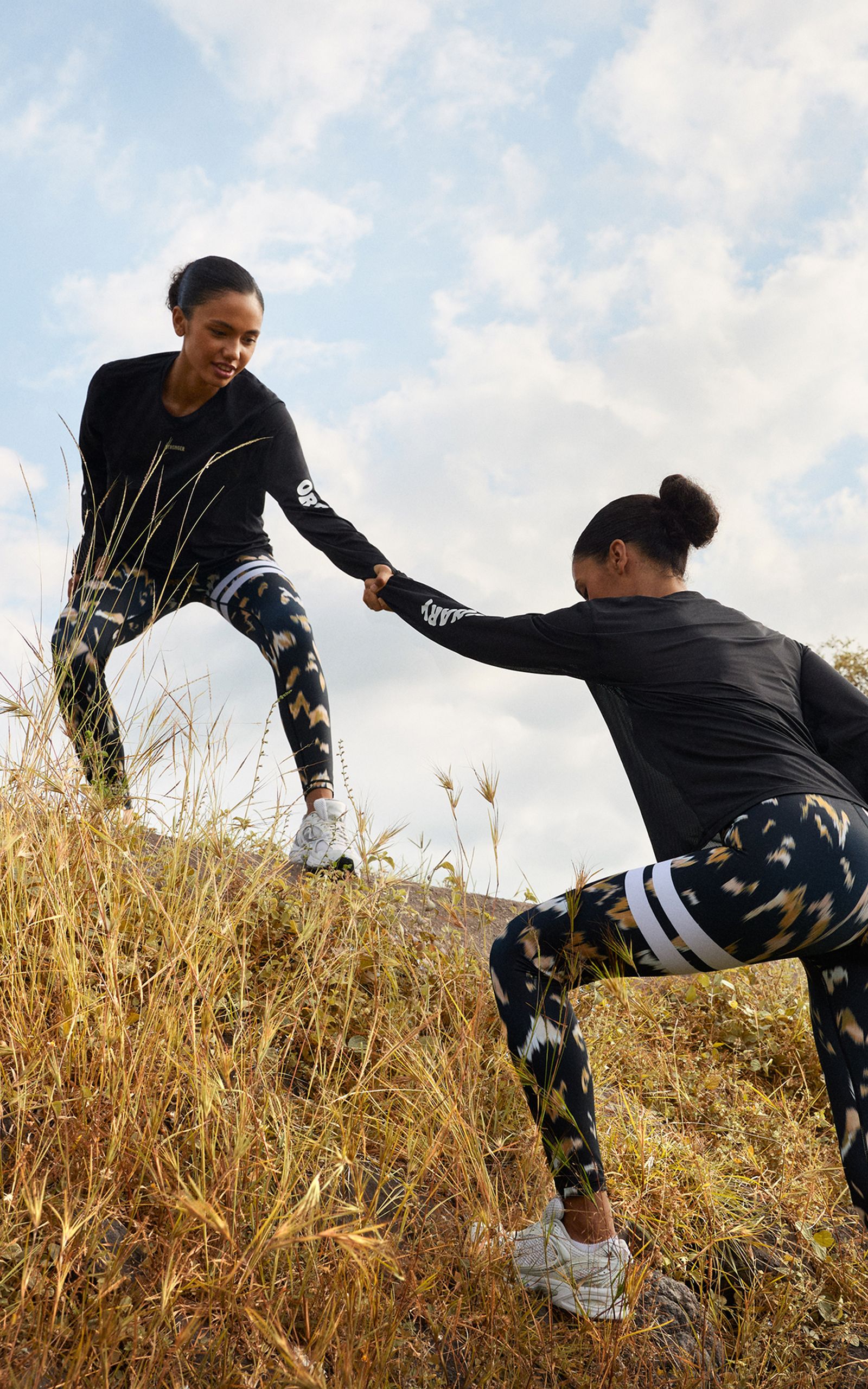 Two women in athletic wear climbing a grassy hill, one helping the other up under a partly cloudy sky.