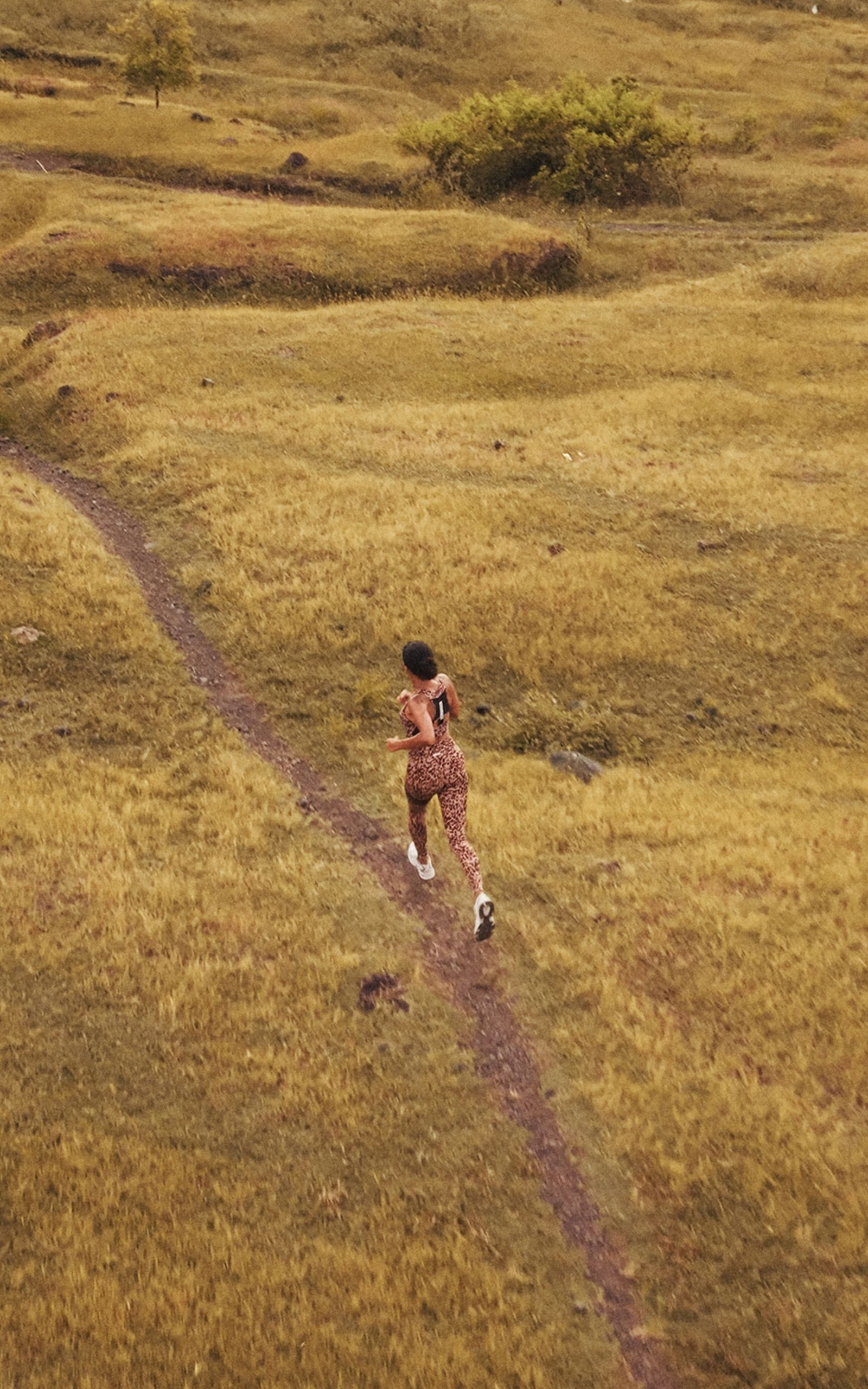 Runner in leopard-print outfit jogging along a narrow dirt path through a grassy field, surrounded by gentle hills.