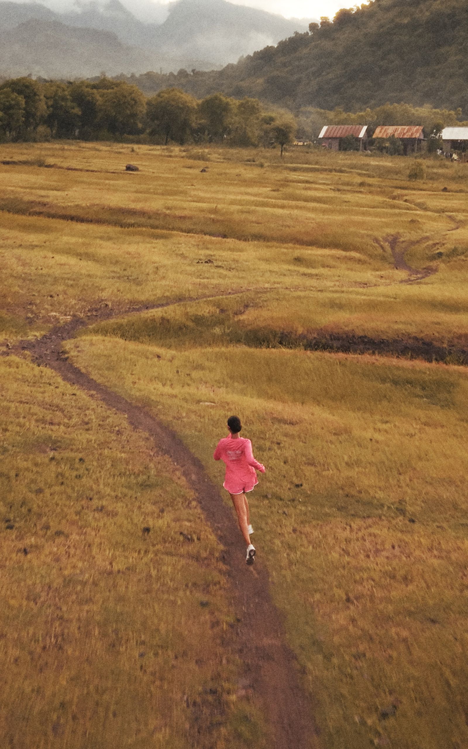 A person in a pink jacket runs on a winding path through a grassy field with distant mountains and a few houses in the background.