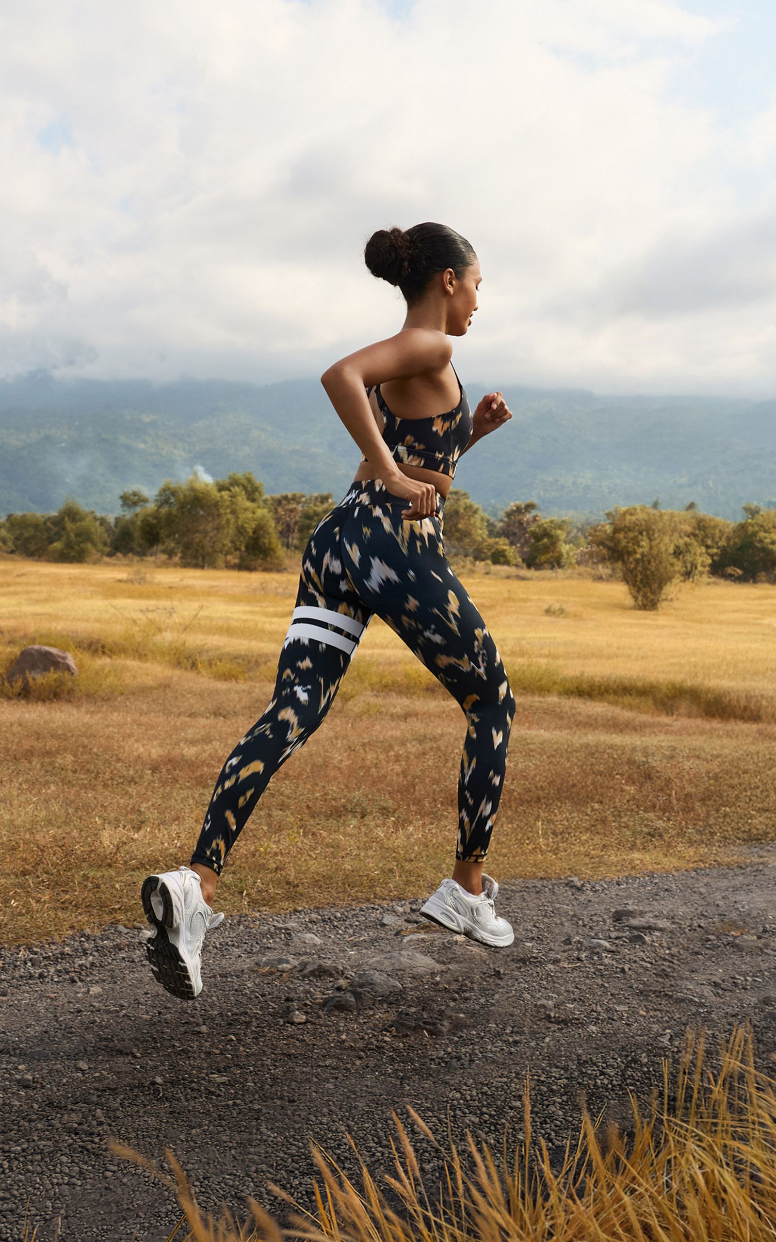 Woman running on a rural path, wearing a patterned sports outfit, with mountains and fields in the background.