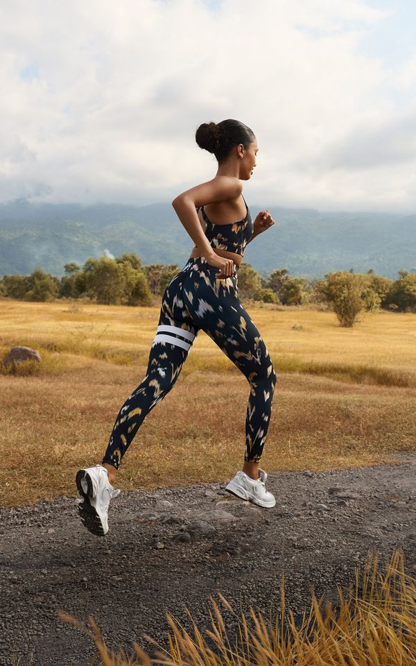 Woman running on a rural path, wearing a patterned sports outfit, with mountains and fields in the background.
