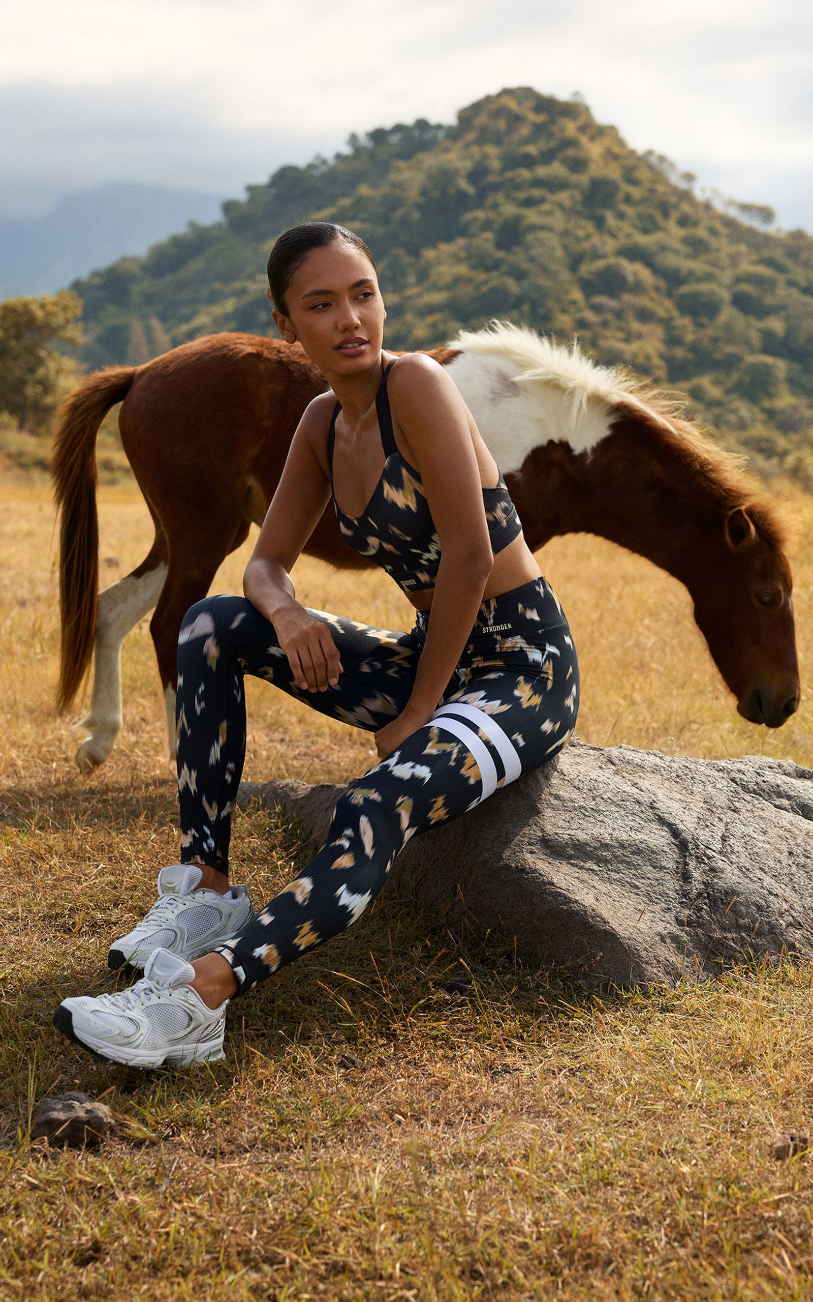A woman in patterned activewear sits on a rock in a grassy field with a brown horse grazing nearby, against a mountainous background.