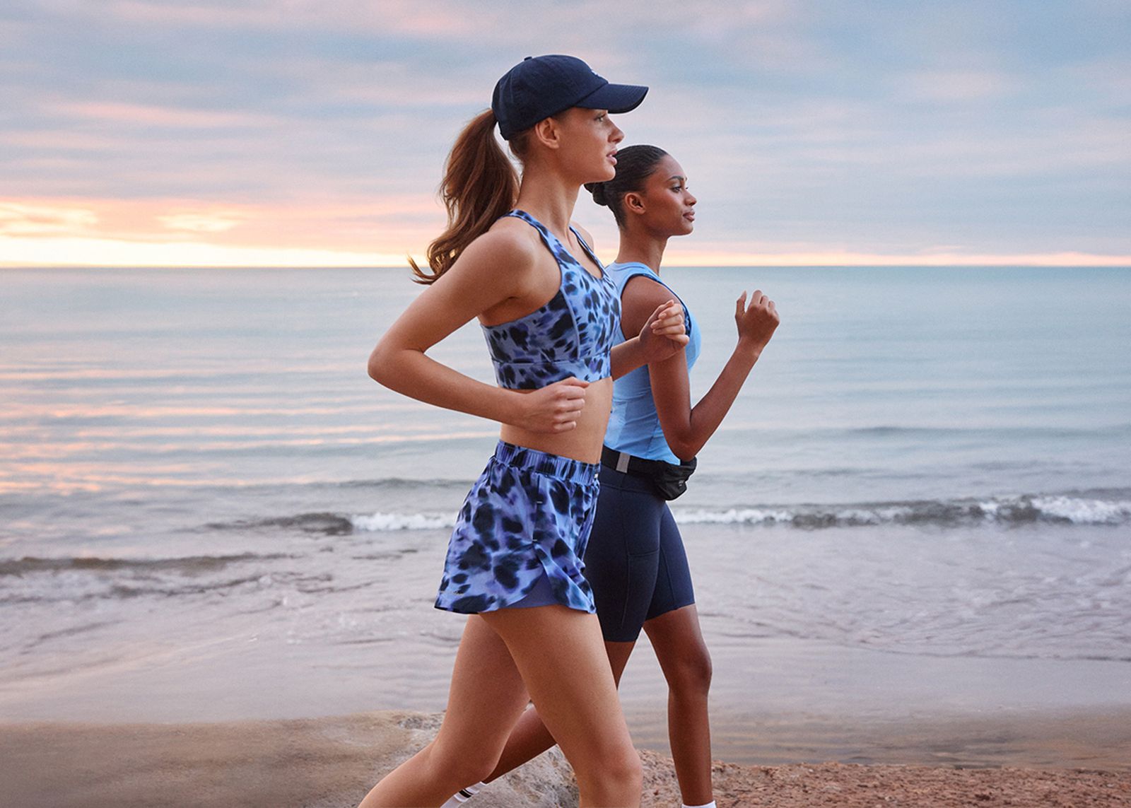 Two women jogging on a beach at sunrise, wearing sportswear and a cap, with a calm ocean in the background.