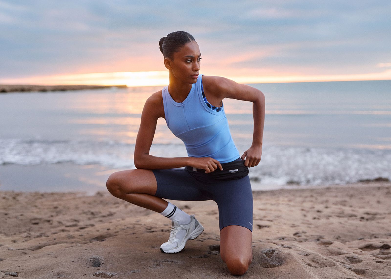 A woman in athletic wear kneels on a beach at sunset, adjusting a pouch around her waist as waves lap the shore.