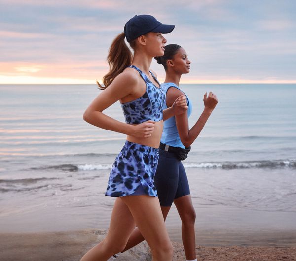 Two women jog along a beach at sunrise, wearing athletic outfits. The sky is colorful, and the ocean waves gently hit the shore.