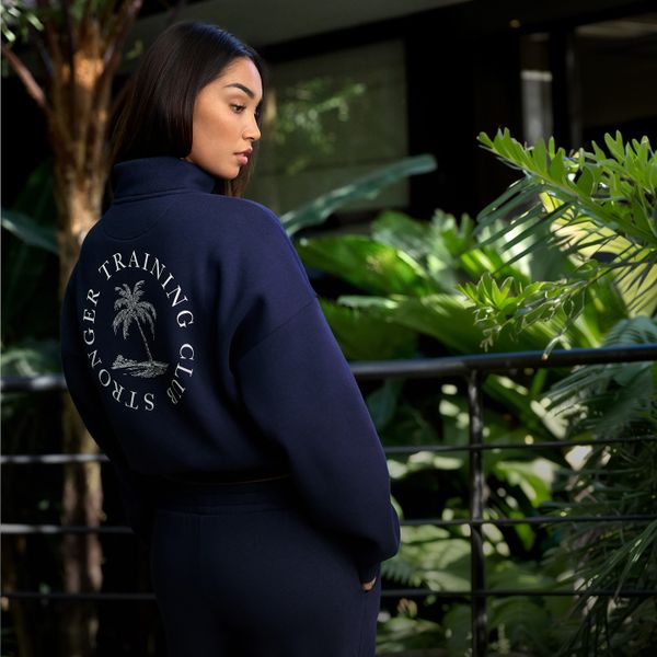 Woman in a navy track jacket with "Stronger Training Club" and a palm tree logo on the back, standing in a lush, green setting.