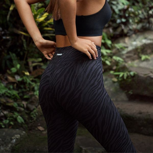 Person wearing a black sports bra and zebra-patterned leggings, standing outdoors with hand on hip and lush greenery in the background.