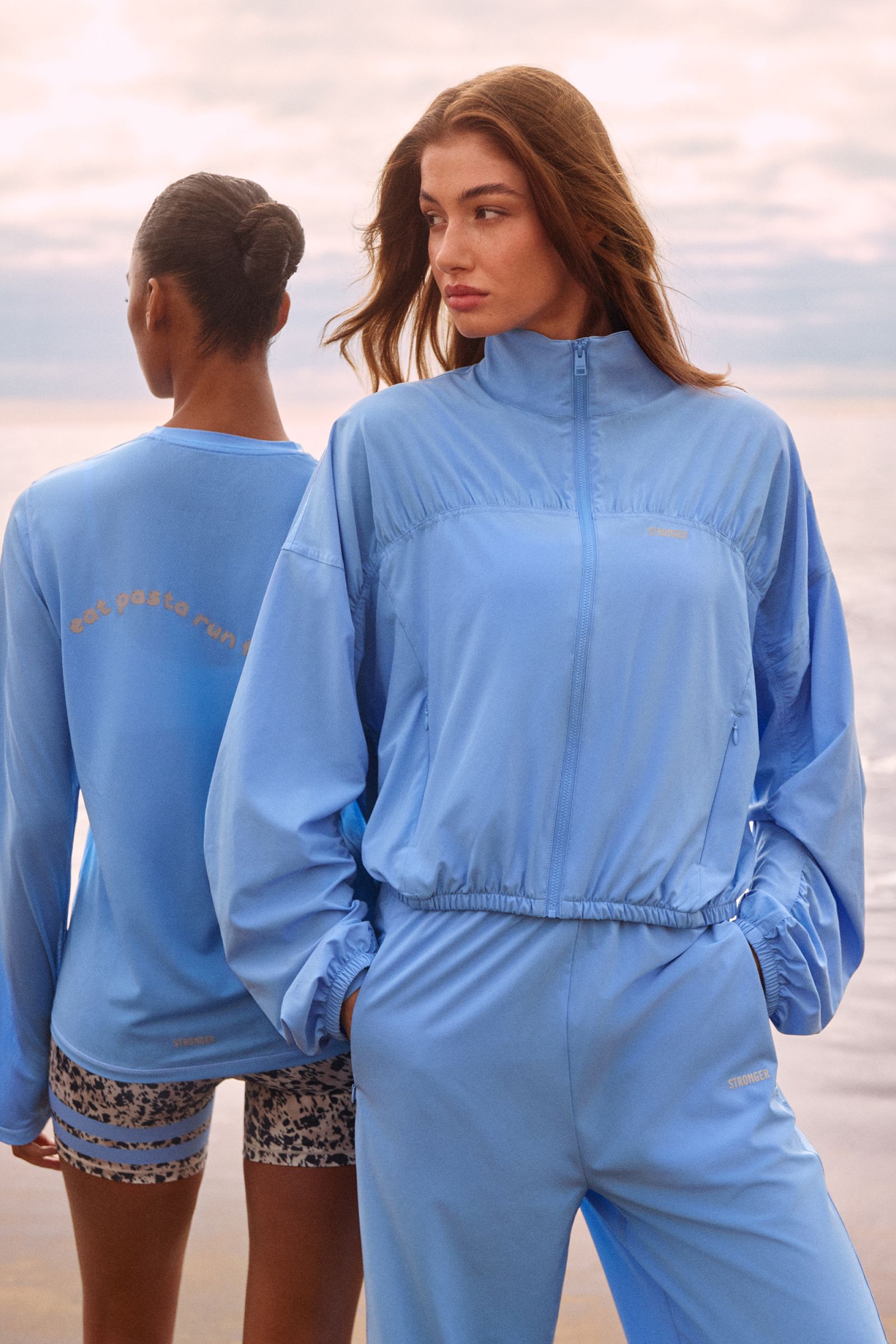 Two women in blue athletic wear stand by the ocean. One faces forward; the other has her back turned, showcasing "eat pasta run" text.