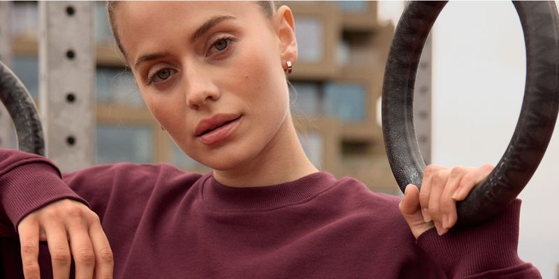Woman in a maroon sweater holding gymnastic rings outdoors, with a focused expression and blurred buildings in the background.