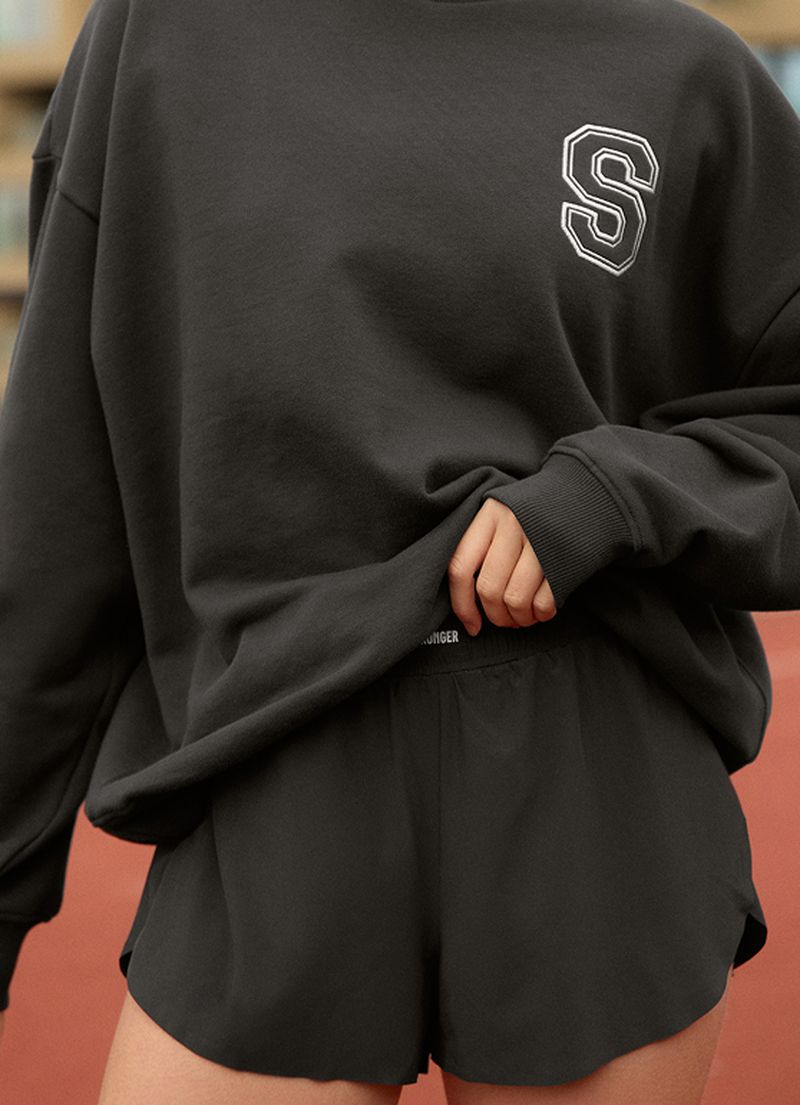 Person in an oversized dark sweatshirt with an "S" and matching shorts, standing on a sports track, holding the sweatshirt hem.