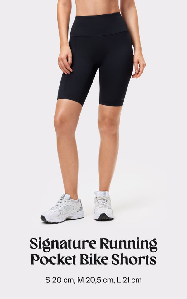 Woman wearing black running pocket bike shorts and white sneakers, shown from midriff down. Text reads: "Signature Running Pocket Bike Shorts."