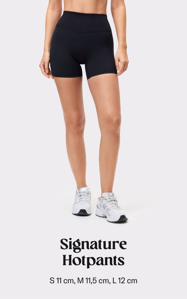 Woman wearing black high-waisted hotpants and white sneakers on a light background, with text "Signature Hotpants" and size details below.