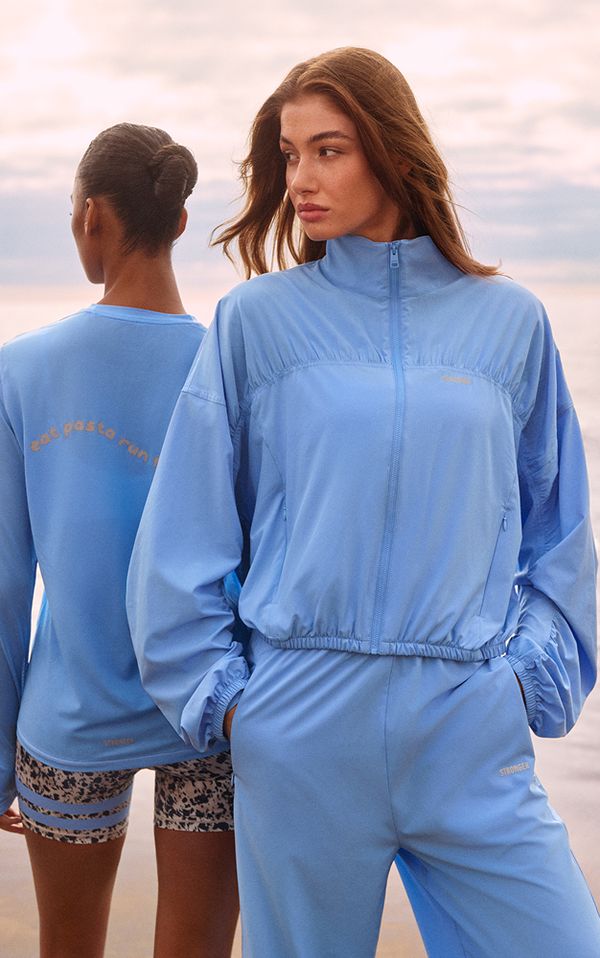 Two women in blue athletic wear, one facing away and the other sideways, on a beach with a cloudy sky backdrop.