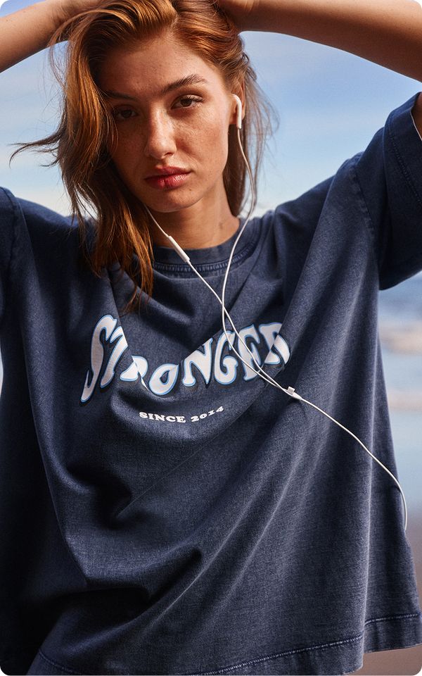 Woman with long red hair wearing a "Stronger" printed t-shirt, earphones in, stands outdoors with arms raised, in a casual pose.