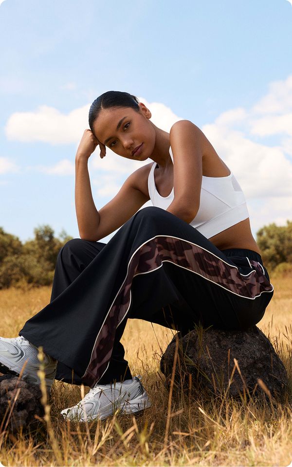 A woman in a white crop top and black pants with stripes sits on a rock in a grassy field, under a blue sky with clouds.