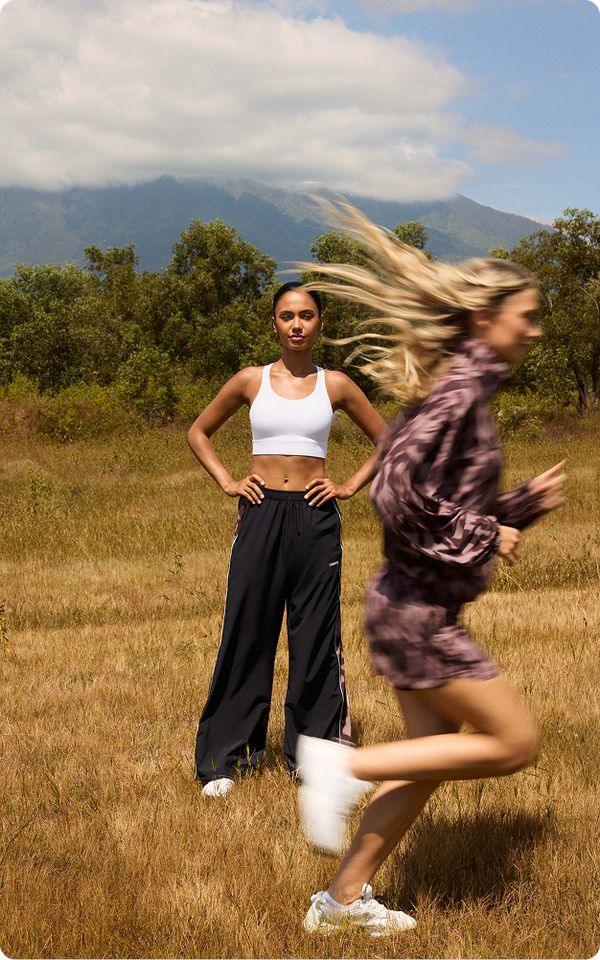 Two women outdoors: one standing in athletic wear, the other running past in a blurred motion, with trees and mountains in the background.
