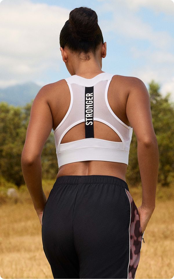 A person in a white sports bra with "STRONGER" on the back stands outdoors, wearing black shorts with a pink pattern, against a natural background.