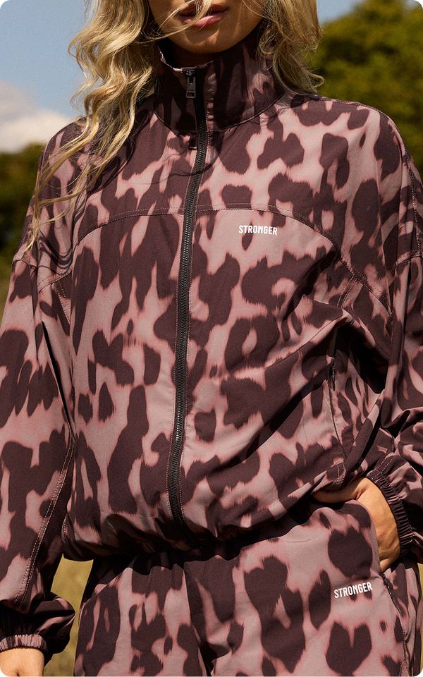 Person in a patterned tracksuit with "STRONGER" text. Light brown and black colors. Outdoors with blurred trees in the background.
