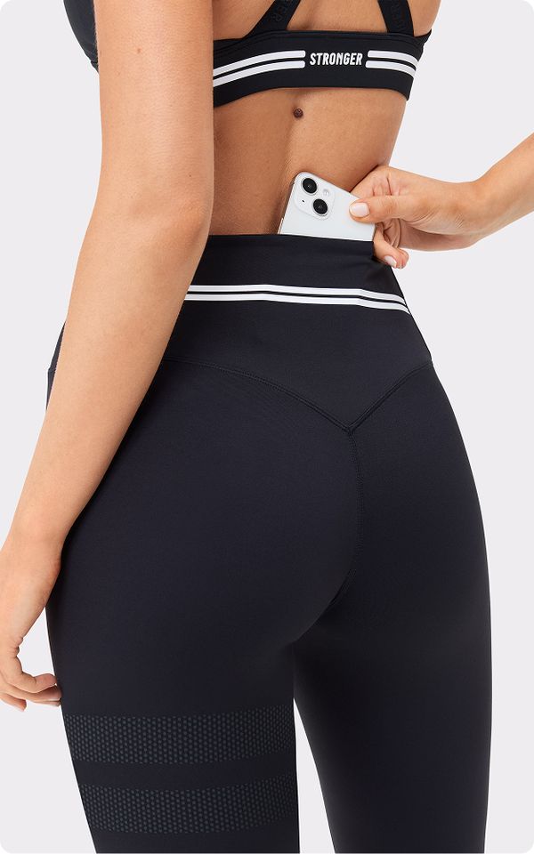 A person wearing black activewear inserts a smartphone into the waistband pocket of their high-waisted leggings.