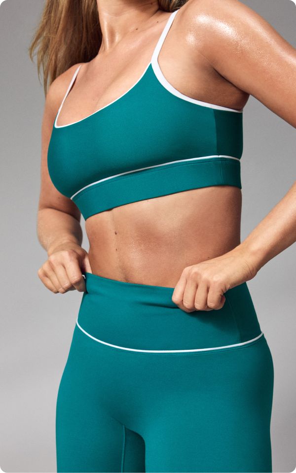 A person wearing teal athletic wear, adjusting high-waisted leggings, showing a fit upper body and light perspiration.
