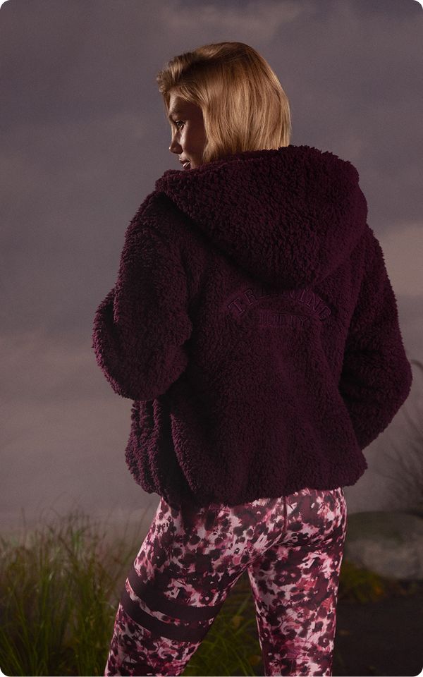 A woman in a fluffy dark purple hoodie stands against a cloudy sky background, wearing floral-patterned leggings.