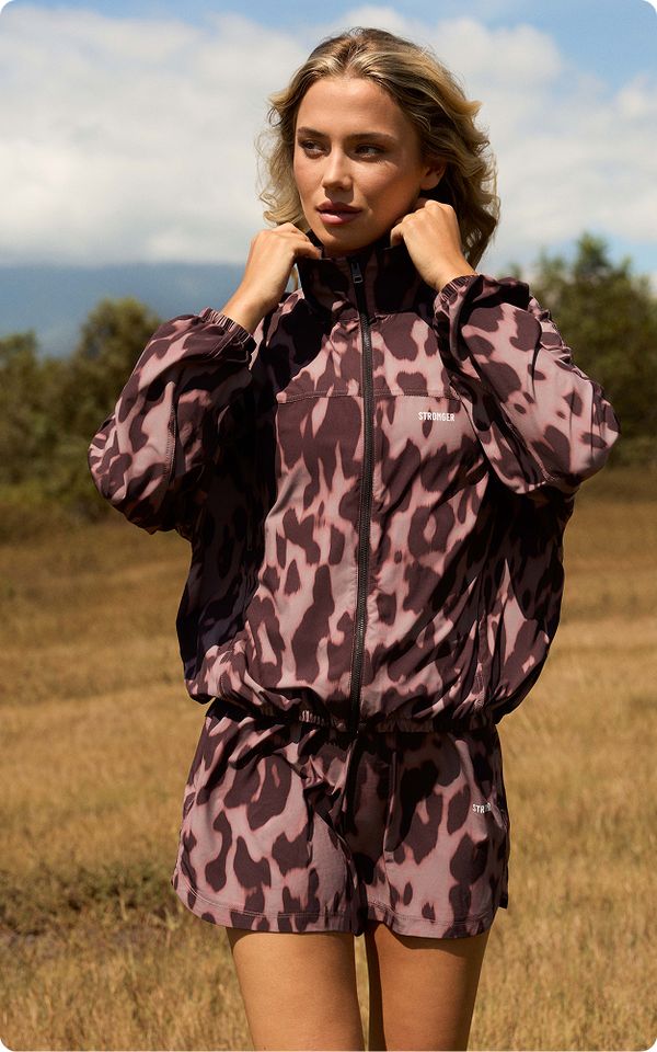 Woman in nature wearing a patterned jacket and shorts, adjusting collar. Brown grass and trees in the background under a cloudy sky.