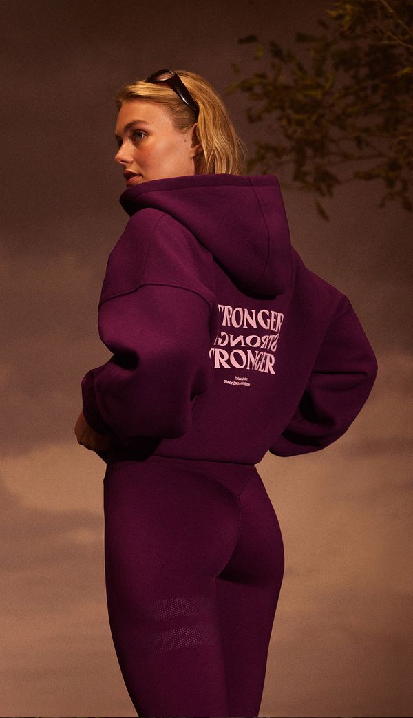A woman wearing a purple hoodie and leggings with "STRONGER" printed on the back, posing against a blurred outdoor background.