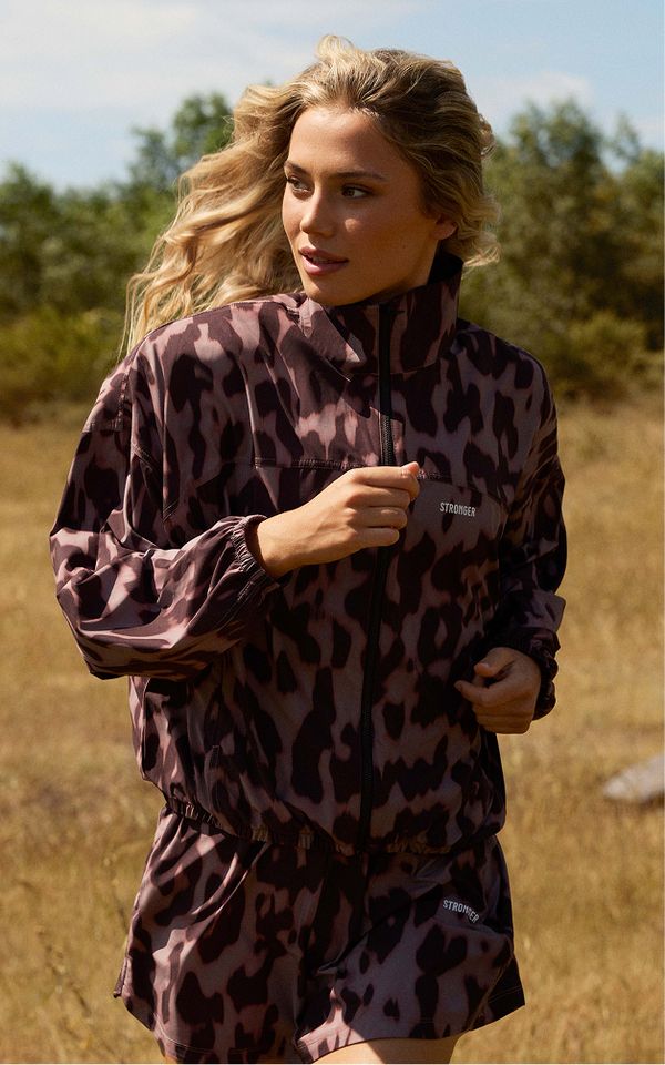 Woman running outdoors in a leopard-print jacket and shorts, with a natural background of trees and grassy fields.