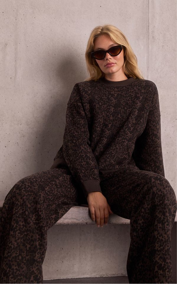 Woman with blonde hair wearing sunglasses and a dark leopard-print outfit, sitting against a concrete wall.