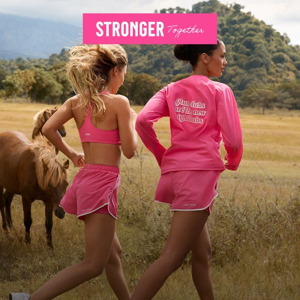 Two women jogging in a field wearing matching pink athletic outfits, with small horses grazing nearby and hills in the background.