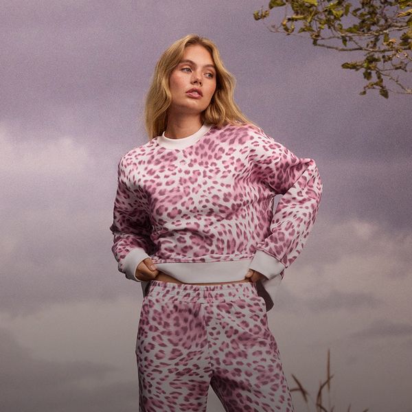 Woman wearing a pink and white leopard print tracksuit, standing outdoors against a cloudy sky, looking to the side.