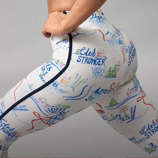 Person wearing white leggings with colorful ski-themed illustrations, standing in a side stretch pose.