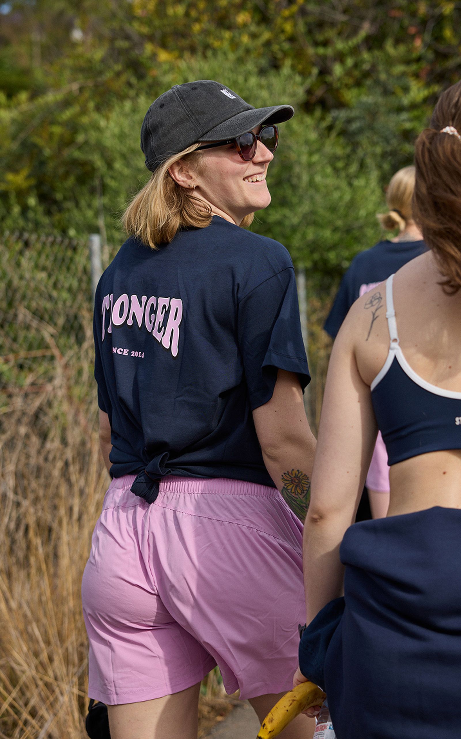Person smiling, wearing a cap, sunglasses, navy "STØNGER" t-shirt, and pink shorts. Walking outdoors holding a banana.