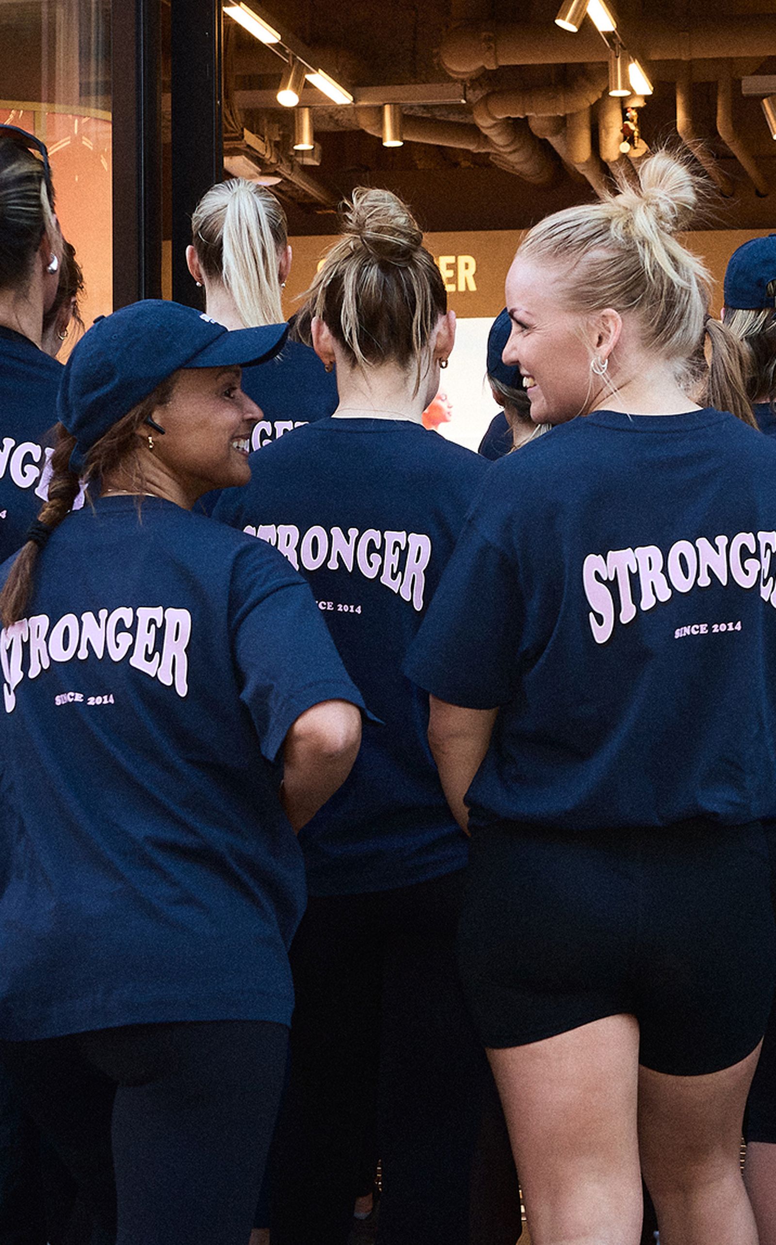 Women in matching navy shirts with "STRONGER" printed on the back, standing in a group, smiling and interacting.