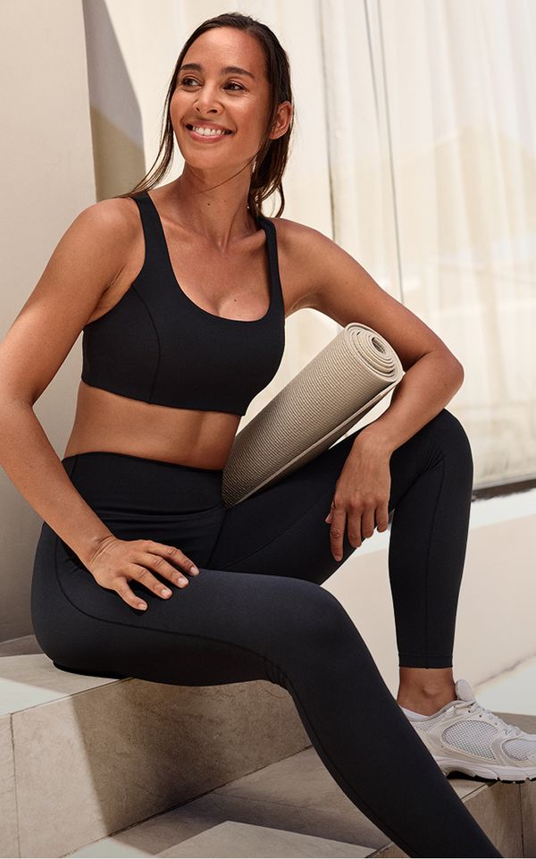 Woman in athletic wear sitting on steps, smiling, holding a yoga mat, wearing a black sports bra and leggings with white sneakers.
