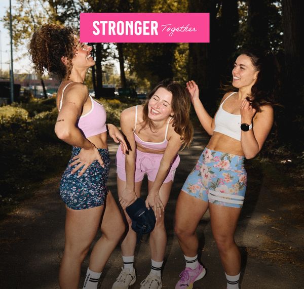 Three women in activewear laughing together on a sunlit path, with trees in the background. Text above reads "STRONGER Together."