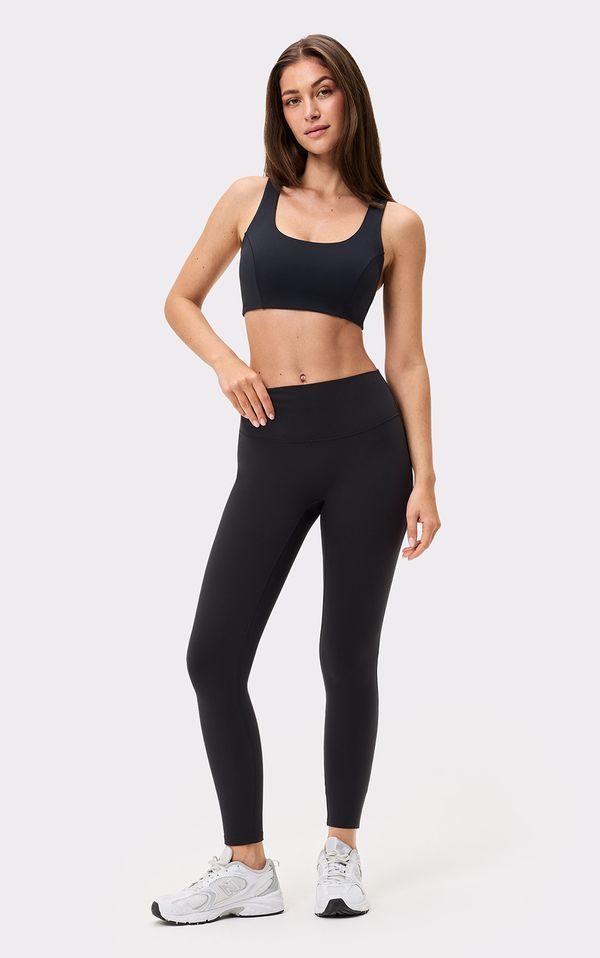 Woman in a black sports bra and leggings, wearing white sneakers, standing against a plain light background.