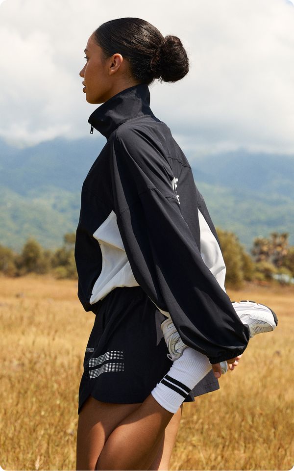 A person stretches in a field, wearing a black and white sports jacket and shorts, with mountains and cloudy sky in the background.