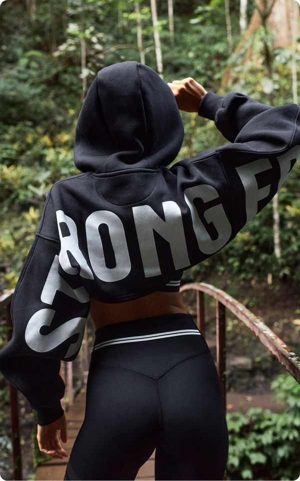 A person wearing a black hoodie with bold white text "STRONGER" stands on a bridge in a lush green forest, looking ahead.