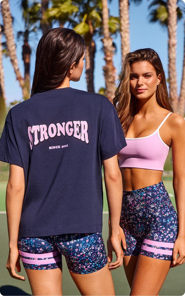 Two women in athletic wear on a sunny day; one faces the camera in a pink top, the other shows a "STRONGER" T-shirt logo. Palms in the background.